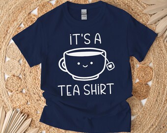 May include: Navy blue t-shirt with the text "IT'S A TEA SHIRT" and a smiling teacup graphic in white. The teacup has a tea bag attached. The shirt is a casual wear item.