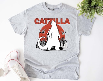 Może przedstawiać: Szara koszulka z nadrukiem białego potwora-kota. Potwór-kot stoi przed czerwonym słońcem i panoramą miasta. Tekst "CATZULLA" jest nadrukowany na czerwono nad grafiką.