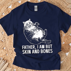 May include: A navy blue short-sleeve t-shirt featuring a white graphic of an overweight cat lying on its back with a small bowl of food. Below the cat, the text reads: FATHER, I AM BUT SKIN AND BONES. The shirt has a crew neckline and a standard fit.