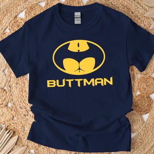 Op de afbeelding: Marineblauw t-shirt met een gele grafische afbeelding. Het ontwerp is een parodie op het Batman-logo, met een vleermuisvormig silhouet en het woord "BUTTMAN" in gele letters. Het t-shirt is gemaakt van zacht materiaal.