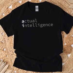 Może przedstawiać: Czarny t-shirt z białym napisem "actual intelligence". Koszulka ma okrągły dekolt i jest wykonana z miękkiego materiału. Tekst jest wyśrodkowany na przodzie koszulki.