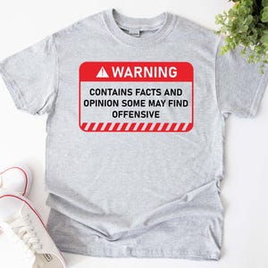 Könnte beinhalten: Ein hellgraues T-Shirt mit einem rot-weißen Warnschild. Das Schild lautet "WARNING CONTAINS FACTS AND OPINION SOME MAY FIND OFFENSIVE". Weiße Turnschuhe mit roter Umrandung sind unten links zu sehen.