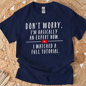 May include: A navy blue short-sleeve t-shirt featuring white text that reads: DON'T WORRY, I'M BASICALLY AN EXPERT NOW. A small red YouTube play button icon is centered between the lines, followed by the text: I WATCHED A FULL TUTORIAL.