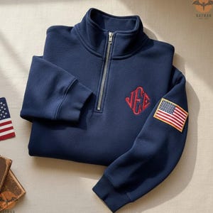 May include: Navy blue quarter-zip sweatshirt with a red monogram and an American flag patch on the sleeve. The sweatshirt is folded and has a silver zipper. The overall theme is patriotic apparel.