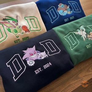 May include: A collection of embroidered sweatshirts in various colors, including tan, navy blue, green, and black. Each sweatshirt features a large "D" design with a different cartoon character and the year it was established. The sweatshirts are displayed on a wooden surface.