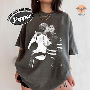 May include: Pepper Comfort Colors t-shirt in charcoal gray featuring a black and white image of two hockey players. One player is kissing the other on the cheek. The shirt also has a Batman logo.