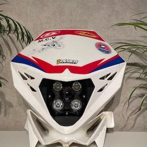 Peut inclure: Phare de moto blanc avec des accents rouges et bleus. Le phare comporte un panneau noir avec quatre lumières rondes. Le haut de l'ensemble porte les inscriptions "M.C.V." et "POWER", ainsi qu'un dessin de moto.