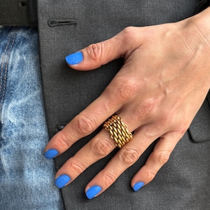 May include: A gold-colored ring with a chain-link design. The ring is worn on a hand with blue nail polish. The background includes a gray blazer and blue jeans.