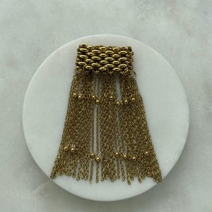 May include: Gold-toned metal tassel pendant. The top section is a rectangular design of small, linked ovals. Below, multiple strands of delicate chains cascade downward, interspersed with small, round beads. The pendant is displayed on a white, circular surface.