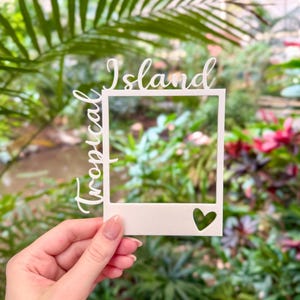 Può includere: Una cornice bianca tagliata al laser con le parole "Tropical Island" in corsivo. La cornice ha un ritaglio a forma di cuore. È tenuta davanti a un lussureggiante sfondo verde.