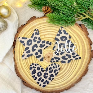 Könnte beinhalten: Weihnachtsschmuck in Stern-, Herz- und Baumform mit einem beigefarbenen und schwarzen Leopardenmuster. Der Baumschmuck trägt den Namen "Jette" in weißer Schrift. Die Ornamente werden auf einer Holzscheibe mit Tannennadeln präsentiert.