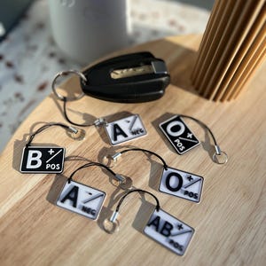 Könnte beinhalten: Ein Satz von sechs Schlüsselanhängern mit verschiedenen Blutgruppen: A+, A-, B+, AB+, O+ und O-. Jeder Schlüsselanhänger ist weiß mit schwarzer Beschriftung und einem roten Plus- oder Minuszeichen.