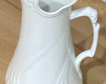 May include: White ceramic pitcher with a decorative design and a handle. The pitcher has a wide mouth and a rounded body.