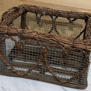 May include: A rectangular, rustic wire basket with a wooden base and frame. The basket is adorned with woven vines and two heart-shaped vine decorations on the front. The basket is designed for decorative storage.