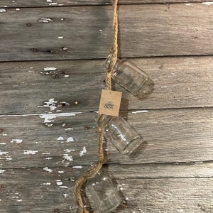 May include: Three clear glass jars hanging from a natural twine rope. The jars are suspended from a wooden wall.