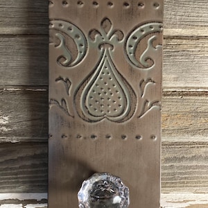 May include: A brown metal wall hook with a decorative embossed design and a clear glass knob. The hook is mounted on a wooden surface.