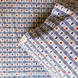 May include: A white and blue checked fabric with a repeating pattern of red apples, blue flowers, and white dots. The fabric has a label that says "ODA NONOT".