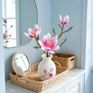 May include: A white crocheted vase with pink floral embroidery holds crocheted magnolia flowers with pink and white petals. The vase sits on a woven tray with a small round mirror and a wicker basket.