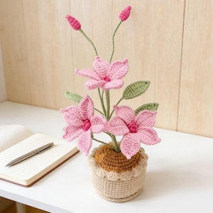 May include: A handcrafted crochet floral display with pink blooms, green foliage, and buds. The flowers are presented in a beige pot with a scalloped trim. The arrangement is on a white surface, with a notebook and pen visible.