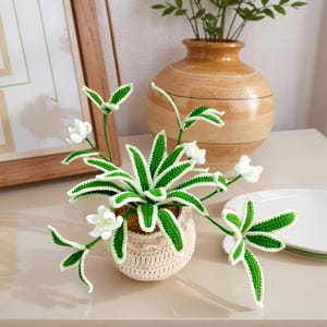 以下が含まれることがあります： 手作りのかぎ針編みの植物アレンジメント。クリーム色の小さなポットにレースのトリムが施されています。ポットには、白い花が付いた緑と白のかぎ針編みの植物が入っています。木製の花瓶と額入りのアートワークが背景にあります。