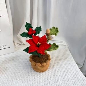 May include: A crocheted poinsettia plant in a tan pot with a brown rim. The plant features a bright red poinsettia flower with yellow centers, green holly leaves with red berries, and two small acorns with tan and white caps attached to green leaves.