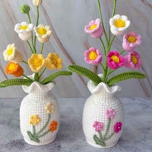 以下が含まれることがあります： 2つの白いかぎ針編みの花瓶。それぞれカラフルなかぎ針編みの花で満たされています。1つの花瓶には黄色、オレンジ、白の花があります。もう1つはピンクと白の花です。緑の葉と茎が花の配置を完成させています。