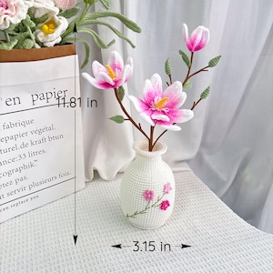 May include: A crocheted white vase with pink floral embroidery holds pink and white crocheted magnolia flowers. The vase is 3.15 inches wide and the flowers are displayed on a table. The height of the paper bag is 11.81 inches.