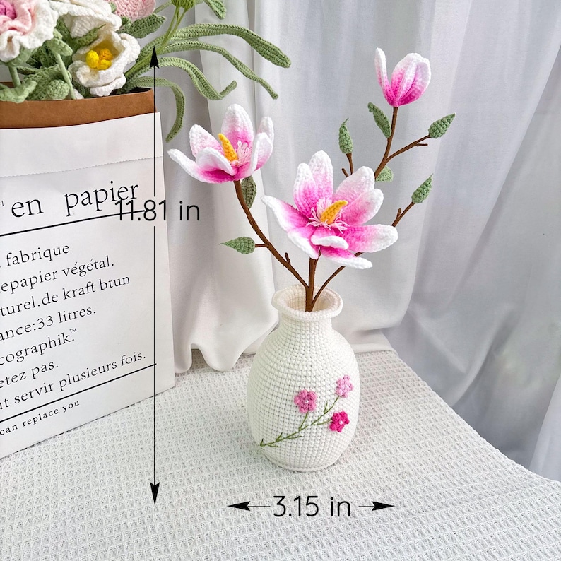 May include: A crocheted white vase with pink floral embroidery holds pink and white crocheted magnolia flowers. The vase is 3.15 inches wide and the flowers are displayed on a table. The height of the paper bag is 11.81 inches.