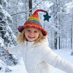 Peut inclure: Une jeune personne porte un chapeau crocheté aux couleurs de l'arc-en-ciel avec une étoile, souriant dans une forêt enneigée. Le chapeau a un bord à volants et un sommet incurvé. La personne porte un manteau d'hiver blanc.