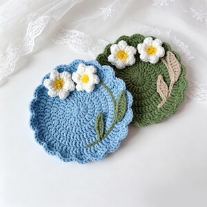 May include: Two handmade crochet coasters. One is light blue with white and yellow flower accents and green leaves. The other is green with white and yellow flower accents and tan leaf accents. Both have a scalloped edge.
