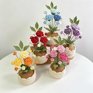 Puede incluir: Una colección de cinco arreglos florales de ganchillo en pequeñas macetas redondas de color crema con tapas marrones. Cada arreglo presenta hojas verdes y flores de varios colores, incluyendo flores amarillas, naranjas, rojas, azules, moradas y rosas.