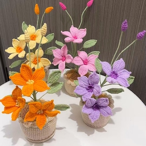 Pode incluir: Uma coleção de flores de crochê feitas à mão em várias cores, incluindo amarelo, laranja, rosa e roxo. Cada flor está em um pequeno cesto tecido. Folhas e caules verdes adicionam realismo aos arranjos florais.