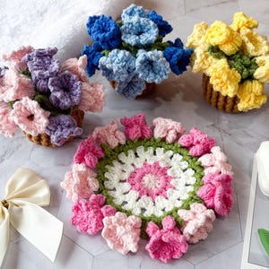 Könnte beinhalten: Handgefertigte Häkelblumenarrangements in kleinen gewebten Körben. Die Blumen sind in den Farbtönen Rosa, Lila, Blau und Gelb gehalten. Ein gehäkelter Untersetzer mit rosa Blüten und einem weißen Zentrum ist ebenfalls vorhanden.