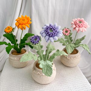 Op de afbeelding: Drie gehaakte bloemstukken in crèmekleurige potten. De bloemen zijn in tinten oranje, paars en roze, met groene bladeren en stelen. De potten hebben een decoratieve kanten rand. De achtergrond is witte stof.