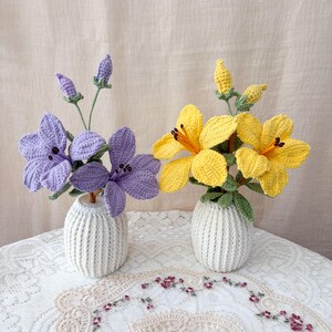 May include: Two crochet flower arrangements. One has purple flowers, the other yellow. Each is in a small, white, ribbed vase. The flowers have green stems and leaves. The background is a neutral colour.