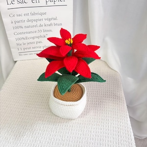 Può includere: Una pianta di poinsettia all'uncinetto in un vaso bianco. La pianta presenta petali rosso vivo, foglie verdi e un centro giallo. Il vaso è bianco con interno marrone e la pianta poggia su una superficie bianca.
