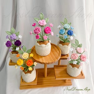 May include: A collection of handmade crochet flower arrangements in small woven pots. The flowers are in various colors, including pink, purple, yellow, and blue, with green leaves. The arrangements are displayed on wooden stands.