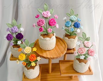 May include: A display of handmade crochet flower arrangements in small woven pots. The flowers are in various colors, including pink, blue, yellow, and purple, with green leaves. The arrangements sit on wooden tiered stands. The text "Nest Dream Shop" is visible.