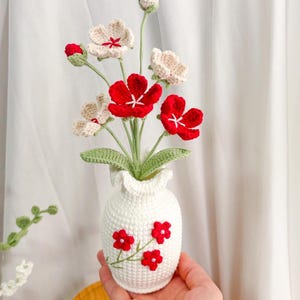 Op de afbeelding: Een handgemaakt gehaakt bloemstuk in een witte vaas. De bloemen zijn rood en gebroken wit met groene stelen en bladeren. De vaas is versierd met rode bloemen. Een charmant, handgemaakt woondecoratie-item.