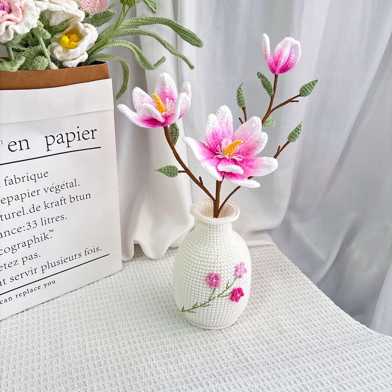 May include: A white crocheted vase with pink embroidered flowers holds a branch of pink and white crocheted flowers. The flowers have yellow centers and green leaves. The vase is on a white surface.