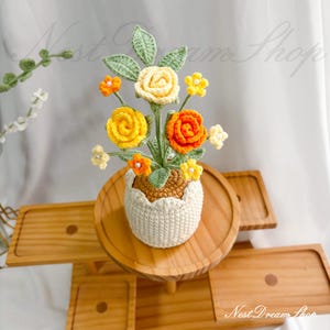 Könnte beinhalten: Ein gehäkeltes Blumenarrangement mit gelben, orangen und cremefarbenen Rosen sowie kleinen Blüten und grünen Blättern. Die Blumen stecken in einem braun-cremefarbenen Häkeltopf, der wie eine zerbrochene, texturierte Eierschale geformt ist.