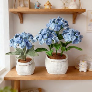 Puede incluir: Dos plantas de hortensias de ganchillo en macetas blancas sobre una estantería de madera. Las flores azules tienen un diseño en capas con pequeñas cuentas blancas. Las macetas tienen un aspecto texturizado y tejido, y las hojas son verdes.