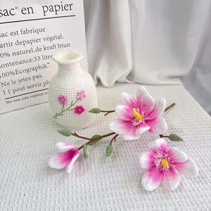 May include: A white crocheted vase with pink embroidered flowers and three crocheted pink and white magnolia flowers with yellow centers. The flowers are on brown stems with green leaves. The background includes a white paper bag.