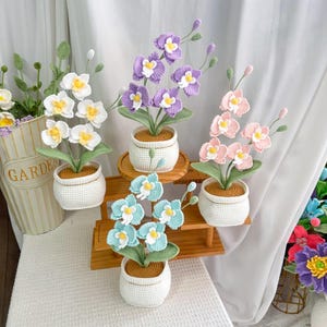 Op de afbeelding: Gehaakte orchideeënplanten in witte potten. De bloemen zijn in tinten wit, paars, roze en blauw met gele centra. De planten staan op een houten plank en een wit oppervlak. De achtergrond omvat een wit gordijn en een metalen emmer.