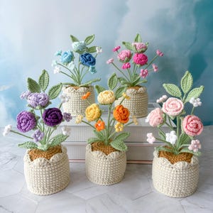 Puede incluir: Cinco arreglos florales de ganchillo en macetas color crema. Las flores son de varios colores, incluyendo azul, rosa, amarillo, naranja y morado. Cada arreglo tiene hojas verdes y está sobre un fondo azul claro.