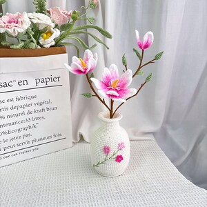 May include: A white crocheted vase with pink embroidered flowers holds a branch of pink and white crocheted magnolia flowers. A paper bag with text in French is in the background, containing more crocheted flowers.