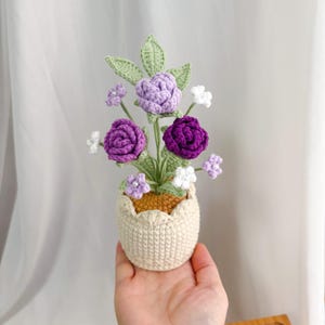 May include: A crocheted floral arrangement featuring purple and white flowers, green leaves, and a beige pot. The flowers are in varying shades of purple, with white accents. The pot has a scalloped edge.