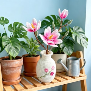 May include: A white crocheted vase with pink floral embroidery holds a branch of crocheted pink and white magnolia flowers. The vase sits on a wooden shelf with potted plants and a small metal watering can.