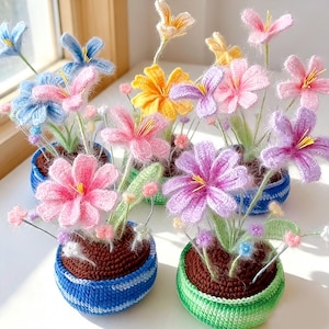 以下が含まれることがあります： 小さな丸い質感のある鉢に入ったかぎ針編みの花のコレクション。花はピンク、紫、青、黄色の鮮やかな花びらと細い緑の茎が特徴です。鉢は青と緑のかぎ針編みで、上部は茶色の土のような質感になっています。