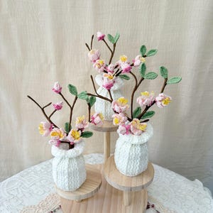 May include: Three crocheted flower arrangements. Each arrangement features a white knitted vase holding branches with pink, yellow, and green crocheted flowers and leaves. The vases sit on small wooden stands.